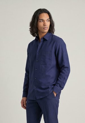 Man met donker haar tot op de schouders, gekleed in een donkerblauw overhemd met lange mouwen en knopen en bijpassende broek, kijkt opzij.