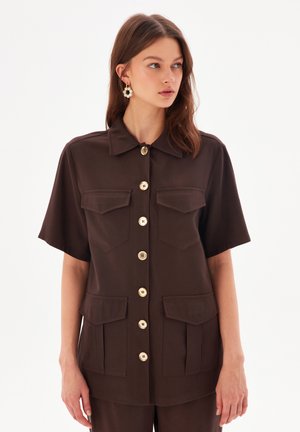 Camisa de tirantes corta marrón con dos bolsillos frontales y botones en tono dorado. Textura suave y un diseño de cuello clásico.