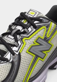 Γκρι και κίτρινο αθλητικό παπούτσι New Balance με μαύρα κορδόνια και πάνω μέρος από δίχτυ, με έμφαση στο λογότυπο "N" στο πλάι.