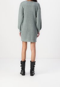 Femme portant une robe en maille gris à manches longues et des bottes noires mi-mollet avec plusieurs boucles, debout contre un fond blanc, vue de dos.