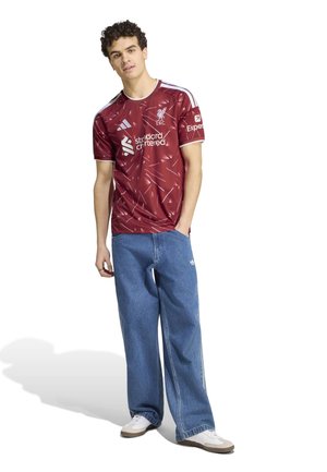 Jeune homme portant un maillot de football rouge du Liverpool FC, un jean large bleu et des baskets blanches, debout avec une main dans la poche.