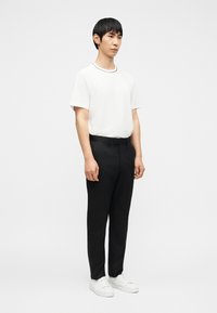 Νεαρός άνδρας όρθιος, φορώντας λευκό κοντομάνικο πουκάμισο, μαύρο παντελόνι και λευκά αθλητικά παπούτσια σε έναν απλό φωτεινό φόντο.