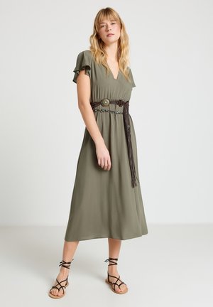 Femme debout portant une robe midi vert olive avec des manches volantées, une ceinture décorative avec des pompons, et des sandales plates noires à lanières.