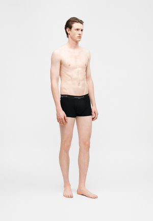 Noor mees heleda nahaga ja pruunide juustega, seisab paljajalu, seljas mustad bokserid vööga "Maison Kitsuné", valgel taustal.