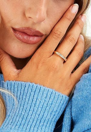 Gold ring with rectangular clear accents, worn on a hand against a blue ribbed sweater. Smooth texture, simple design, elegant.