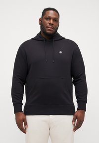 Black hooded sweatshirt with a front pocket and drawstrings. Features a small white logo on the chest and ribbed cuffs and hem.