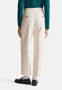 Crèmekleurige broek met een wijduitlopende pasvorm, zichtbare achterzakken en bruine knopen. Ter hoogte van de enkels taps toelopend, gedragen met zwarte schoenen en witte sokken.