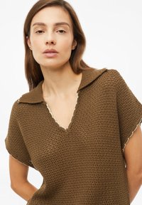 Mujer con cabello castaño liso, vestida con un top de punto marrón texturizado de manga corta con cuello y escote en forma de V sobre fondo blanco.
