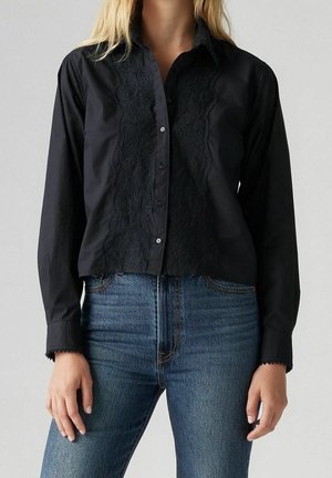 Woman wearing a black button-up shirt with lace detail front paired with high-waisted blue jeans against a plain gray background.