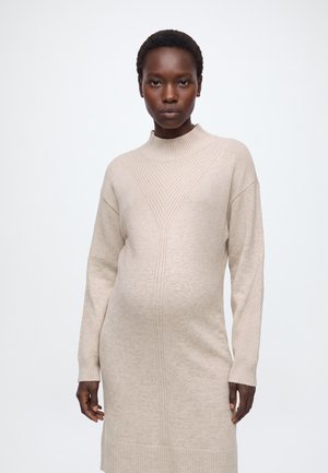 Femme enceinte aux cheveux courts portant une robe beige en maille à manches longues, debout devant un fond blanc uni.