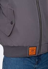 Gray bomber jacket with front zipper, angled flap pocket with button, ribbed hem, orange rectangular label, worn over blue jeans.