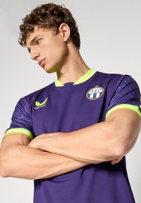 Lila sporttröja med korta ärmar, med neongröna detaljer och ett geometriskt mönster på ärmarna. Inkluderar en logotyp på bröstet.