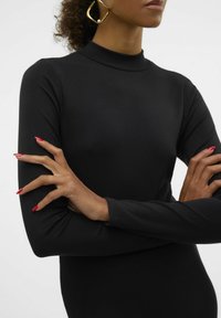 Maglietta nera a coste con maniche lunghe e collo alto. Inquadratura ravvicinata di braccia incrociate, che mette in evidenza il design aderente e la consistenza liscia. Manicure rossa visibile.