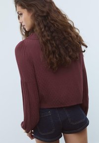 Blouse courte texturée bordeaux à manches longues fluide, associée à un short en denim foncé. La blouse présente un motif subtil.