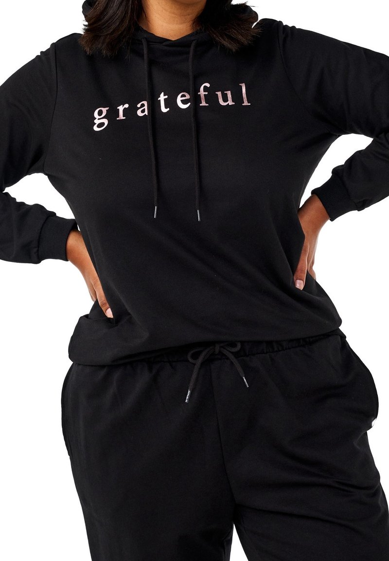 Personne portant un sweat à capuche noir avec le mot « grateful » en lettres rose clair, associé à un pantalon noir à cordon, mains posées sur les hanches.