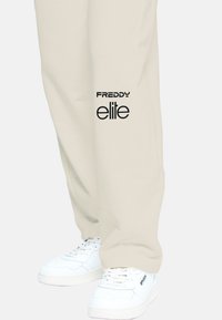 Pantaloni della tuta beige con testo nero "FREDDY" e "elite" sul lato; abbinati a sneaker bianche con dettagli perforati e lacci.