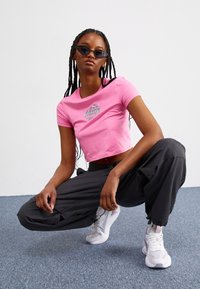 Rosa kort t-shirt med tryckt mönster, kombinerad med löst sittande svarta cargobyxor och vita sneakers. Modellen har flätat hår och solglasögon.