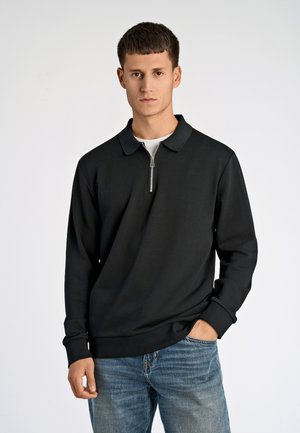Schwarzer Langarmpullover mit einem Reißverschlusskragen, aus einem glatten Stoff. Das Modell trägt ihn zu hellblauen Jeans.