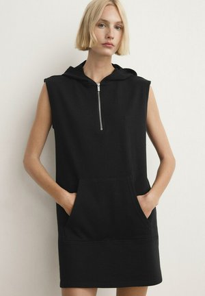 Femme blonde portant une robe noire sans manches à capuche avec une fermeture éclair à l'avant et une poche kangourou, debout les mains dans les poches contre un mur blanc.