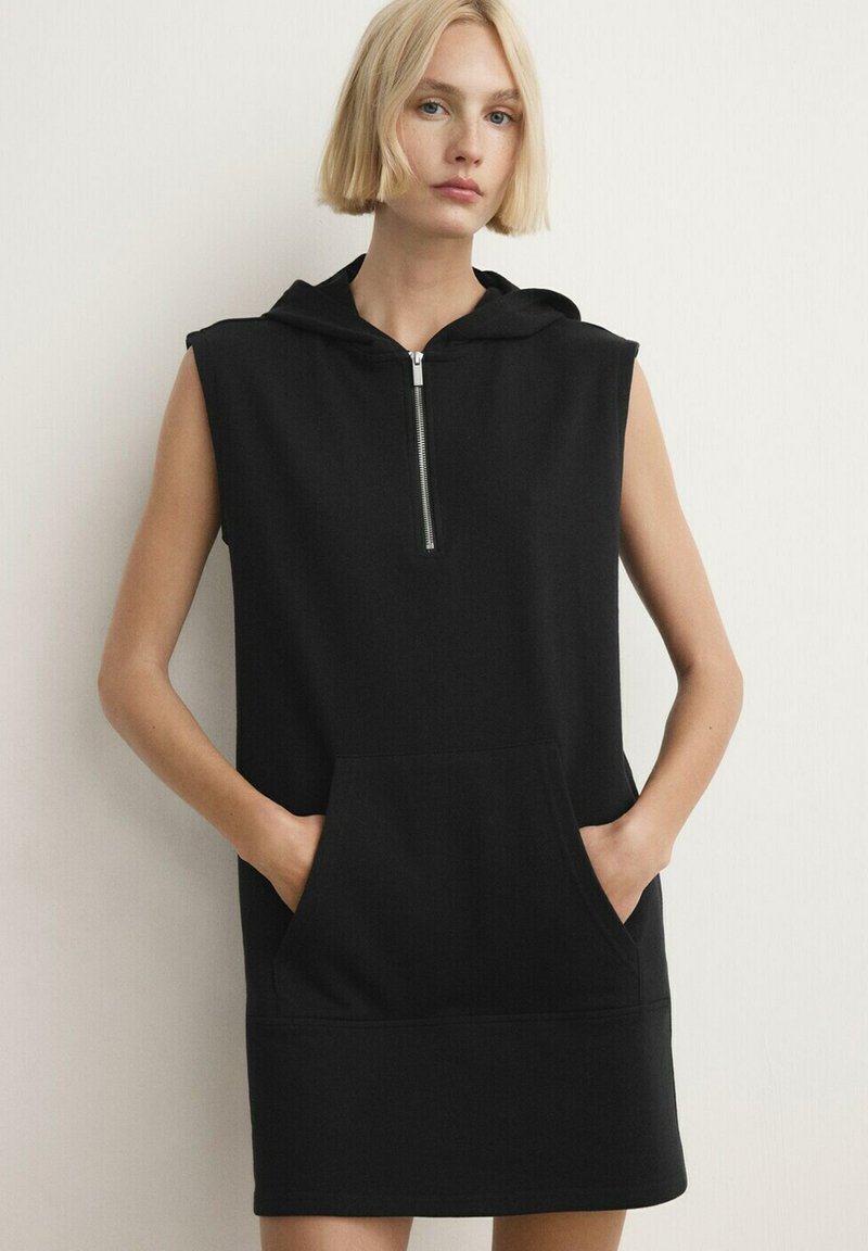 Blonde vrouw draagt een zwarte mouwloze capuchonjurk met ritssluiting aan de voorkant en kangoeroezak, staat met handen in de zakken tegen een witte muur.