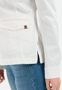 Biała kurtka dżinsowa z przednią kieszenią, bocznymi rozcięciami i widocznym przeszyciem. Pasuje do jasnoniebieskich dżinsów. Gładka faktura, luźny krój.