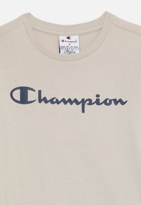 Beige puuvillane T-särk, millel on tume sinine "Champion" logo rinnal ja ümmargune kaelus. Sildikujundus on nähtav krae sees.