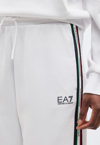 Λευκά παντελόνια φόρμας με το λογότυπο EA7 Emporio Armani και μαύρη, κόκκινη, λευκή και πράσινη ριγέ διακόσμηση, δίπλα σε ένα χέρι με μέτριο σκουρόχρωμο τόνο δέρματος.