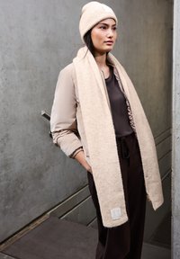 Gorro y bufanda de punto beige combinados con una chaqueta beige con cremallera y pantalones oscuros. Las texturas notables incluyen el canalé de la bufanda y el tejido suave.