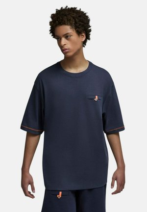 Jeune personne aux cheveux courts et bouclés, vêtue d'une chemise bleu marine ample et d'un short assorti avec de petits détails orange.