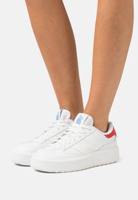 Sneaker bianche con tomaia in pelle liscia, caratterizzate da un accento rosso sul tallone, lacci piatti e una suola in gomma bianca texture. Dettaglio del logo blu.