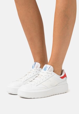 Zapatillas blancas con una parte superior de cuero suave, con un acento rojo en el talón, cordones planos y una suela de goma texturizada blanca. Detalle del logo en azul.
