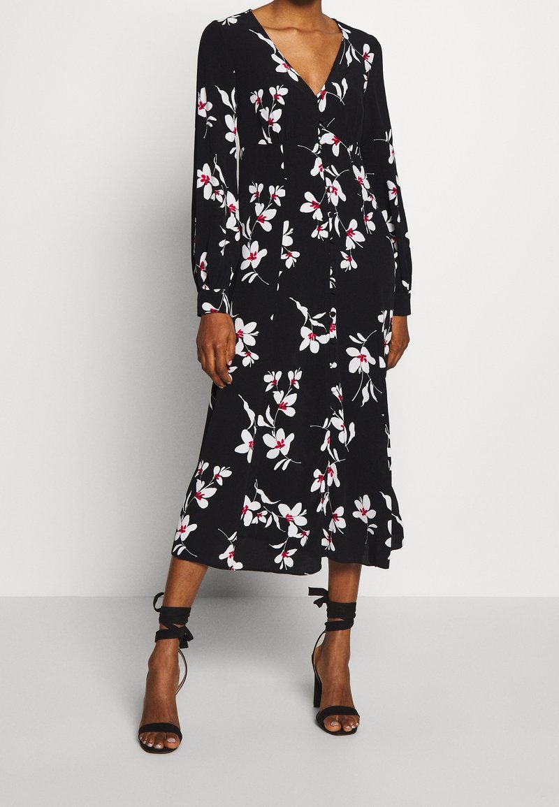 Robe midi noire avec imprimé floral blanc et rouge, manches longues, col en V, portée avec des sandales à talons hauts noires à lacets à la cheville sur une personne debout.