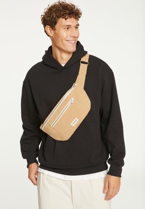 Sac bandoulière en tan fabriqué en tissu durable, doté de deux compartiments zippés, d'un design élégant et d'une sangle réglable, porté en bandoulière.