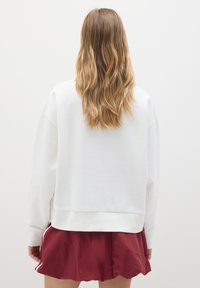Mujer con cabello rubio largo, vistiendo un sudadera blanca y pantalones cortos rojos, de pie frente a una pared blanca lisa.