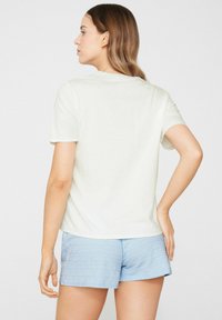 Mujer con cabello largo, con una camiseta blanca de manga corta y pantalones cortos azul claro, de pie con una mano en la cadera, mirando de lado contra un fondo liso.