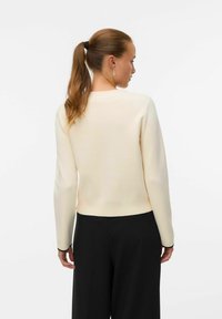 Kremowy dzianinowy sweter z długimi rękawami, okrągłym dekoltem i ściągaczem. W połączeniu z czarnymi spodniami o dopasowanym kroju.