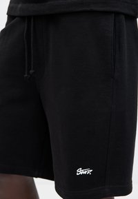 Shorts noirs à cordon de serrage avec poches latérales et petit logo blanc « Swr. » près de l'ourlet, portés par une personne.