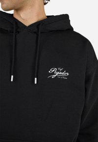 Black hoodie with a drawstring hood. Features embroidered white text "Café Pigador" on the chest. Soft fabric with a casual fit.