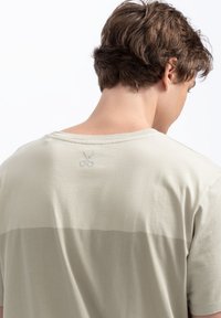 Lichtgroene t-shirt met een horizontale donkerder groene streep, korte mouwen, ronde halslijn en een subtiele schaarlogo dichtbij de achterkant van de halslijn.