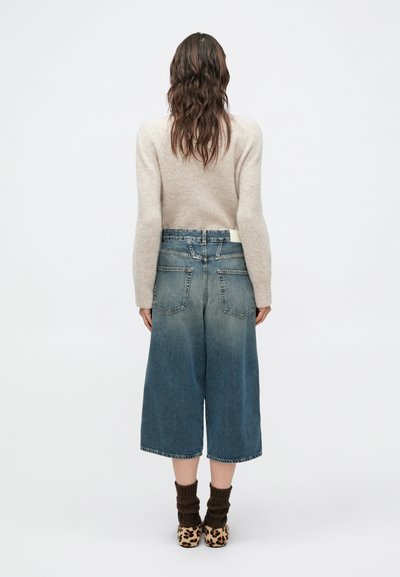 Jean denim bleu à jambes larges avec une finition délavée, taille mi-haute, cinq poches, et un pull beige texturé. Chaussures à imprimé léopard.