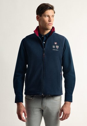 Chaleco de forro polar azul marino con cuello rojo, cremallera frontal, logos bordados y bolsillos laterales. Lleva puesto sobre una camiseta oscura y jeans claros.