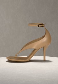 Bēša augstpapēžu sandalete ar potītes siksniņu, smailu purngalu un plānu stiletto papēdi uz gaišas virsmas ar vienkāršu fonu.