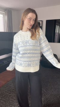 White knit sweater featuring blue geometric patterns, long sleeves, a ribbed hem, and a relaxed fit, paired with black pants.