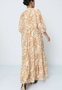 Robe longue et ample avec un motif floral en beige et blanc, dotée de volants superposés et de manches à la longueur du coude. Chaussures à talons hauts noires.