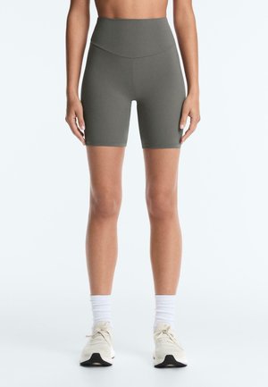 Femme portant un short de cycliste taille haute vert olive, associé à des chaussettes blanches et des baskets écrues, debout devant un fond uni.