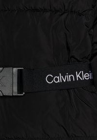 Czarna puchowa kurtka o gładkiej fakturze z szerokim czarnym paskiem i plastikową klamrą, z białym logo „Calvin Klein”.