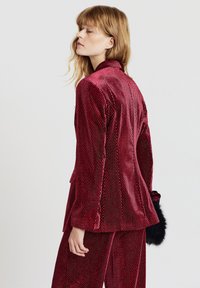 Blazer in velluto rosso con motivo strutturato, vestibilità sartoriale, rever con intaglio e una tasca con patta singola. Dettaglio in pelliccia sul polsino.