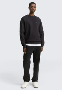 Sort sweatshirt med rund hals og lille logo patch, parret med sorte jeans og hvide sneakers. Glat tekstur, afslappet pasform.