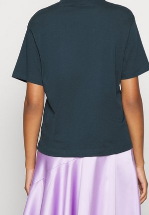 Persoon die een donker turquoise shirt met korte mouwen draagt, gecombineerd met een glanzende lavendelkleurige rok, van achteren gezien tegen een effen achtergrond.