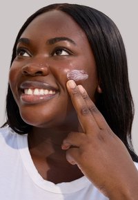 Eine Person mit dunkler Haut lächelt, während sie ein leichtes, cremiges Hautpflegeprodukt auf die Wange aufträgt. Sie trägt ein weißes T-Shirt.